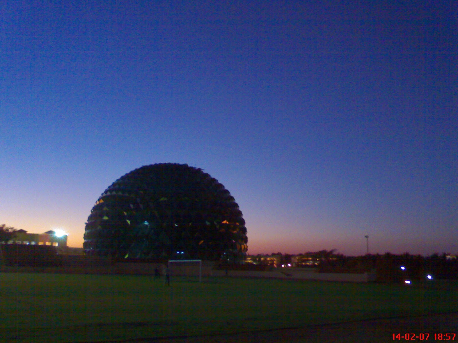 Twilight over a campus in Mysore, India, February 2007. A large geodesic dome silhouetted against a pink and blue sunset sky with a football field in the foreground.